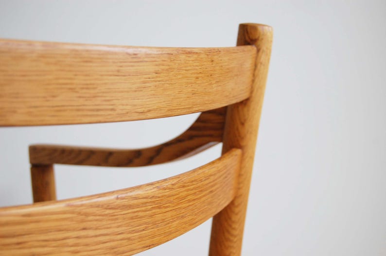May include: Close-up of a wooden chair back. The chair is made of light-colored wood with a visible grain. The design features curved back supports and a vertical post, showcasing a classic furniture style.
