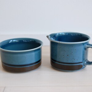 May include: A blue and brown ceramic sugar bowl and creamer set. The bowl and creamer have a speckled glaze and a brown band around the middle.
