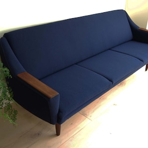 May include: A navy blue mid-century modern sofa with wooden armrests and legs. The three-cushion sofa is placed against a light-colored wall and sits on a light wood floor. A green plant is visible on the left side of the image.