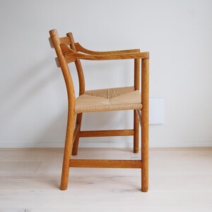 May include: Wooden armchair with a woven seat. The chair has a curved back and armrests. The frame is made of light brown wood. The seat is made of woven natural fibers. The chair is placed against a white wall.