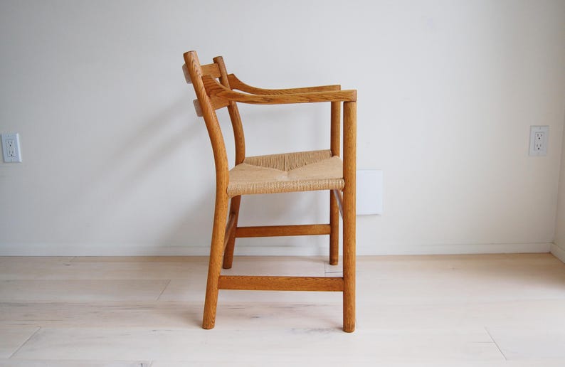 May include: Wooden armchair with a woven seat. The chair has a curved back and armrests. The frame is made of light brown wood. The seat is made of woven natural fibers. The chair is placed against a white wall.