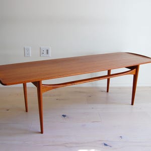 May include: A mid-century modern coffee table with a rectangular top and curved legs. The table is made of wood with a warm, reddish-brown finish. The table measures approximately 60 inches long.