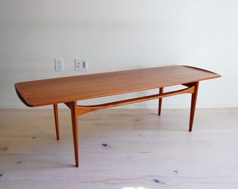 Puede incluir: Una mesa de centro de estilo moderno de mediados de siglo con una tapa rectangular y patas curvas. La mesa es de madera con un acabado marrón rojizo cálido. La mesa mide aproximadamente 152 cm de largo.