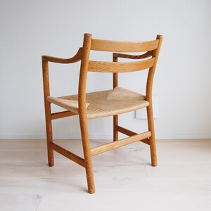 May include: A light-colored wooden armchair with a woven seat. The chair has armrests and a curved backrest. The legs are straight and sturdy. The chair is set against a white wall and light-colored wood floor.