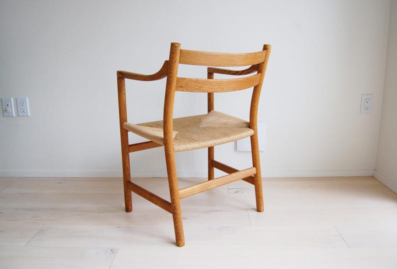 May include: A light-colored wooden armchair with a woven seat. The chair has armrests and a curved backrest. The legs are straight and sturdy. The chair is set against a white wall and light-colored wood floor.