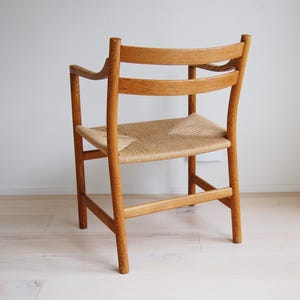 May include: A wooden chair with a woven seat and curved backrest. The chair is made of light-colored wood and has armrests. The seat is made of woven natural fibers. The chair is positioned against a white wall.
