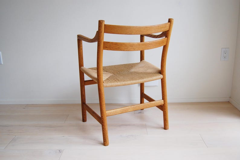 May include: A wooden chair with a woven seat and curved backrest. The chair is made of light-colored wood and has armrests. The seat is made of woven natural fibers. The chair is positioned against a white wall.