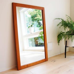 May include: A large, rectangular wooden framed mirror with a light wood finish. The mirror reflects a window with a view of green trees and a potted plant.