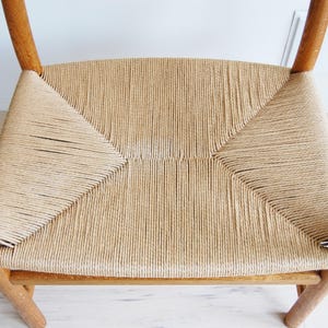 May include: A light-colored wooden chair with a woven seat. The seat is made of tightly woven, light tan rope. The chair's frame is made of light-colored wood. The chair has a classic, minimalist design.
