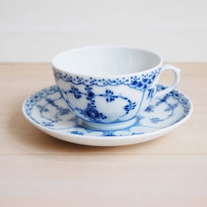 May include: White teacup and saucer set with a blue floral design. The cup has a handle and the saucer is round. The blue pattern decorates the rim of the cup and the edge of the saucer. The set is on a light-colored wooden surface.