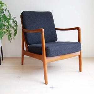 May include: A mid-century modern armchair with a dark gray upholstered seat and back. The chair has a wooden frame with armrests and legs. A potted plant is to the left of the chair.