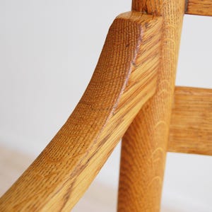 May include: Close-up of a wooden chair with a light brown finish. The chair's armrest and back support are visible, showcasing the wood grain and construction. The chair appears to be made of solid wood.
