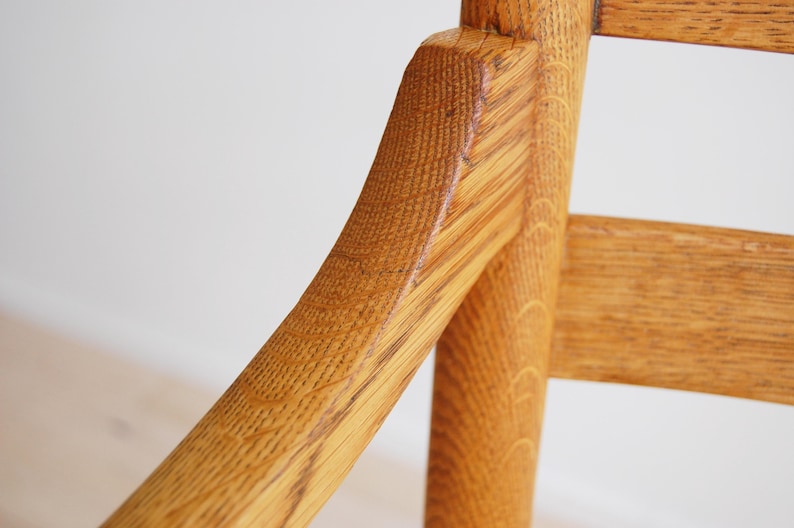 May include: Close-up of a wooden chair with a light brown finish. The chair's armrest and back support are visible, showcasing the wood grain and construction. The chair appears to be made of solid wood.