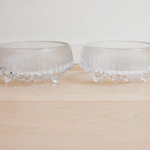 May include: Two clear glass bowls with a textured, vertical ribbed pattern on the exterior sides and a bumpy, irregular base. Each bowl is supported by three small, rounded glass feet, providing a stable foundation for the circular vessels.