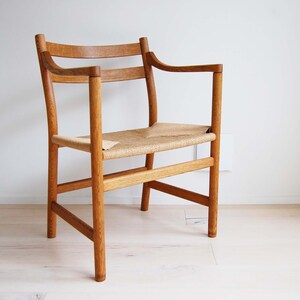 May include: A wooden armchair with a woven seat and armrests. The chair is made of light-colored wood and has a simple, elegant design. The seat is made of woven material. The chair is placed against a white wall.