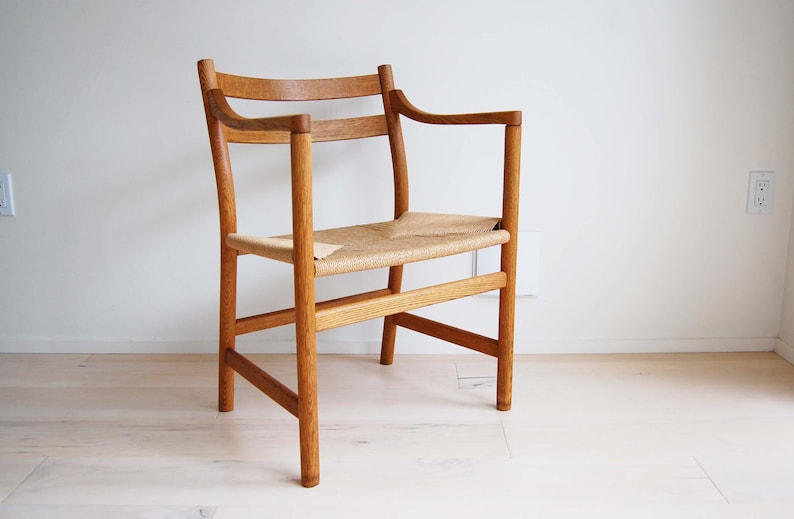 May include: A wooden armchair with a woven seat and armrests. The chair is made of light-colored wood and has a simple, elegant design. The seat is made of woven material. The chair is placed against a white wall.