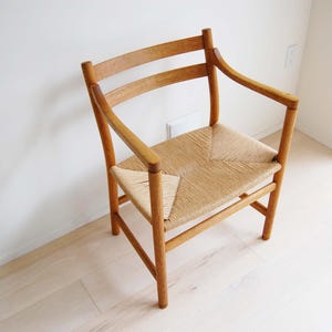 May include: A wooden chair with a woven seat and curved back. The chair has armrests and a light brown finish. The seat is made of woven straw or a similar material. The chair is positioned in a room with light-colored walls and flooring.