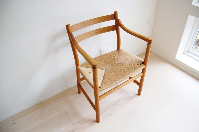 May include: A wooden chair with a woven seat and curved back. The chair has armrests and a light brown finish. The seat is made of woven straw or a similar material. The chair is positioned in a room with light-colored walls and flooring.