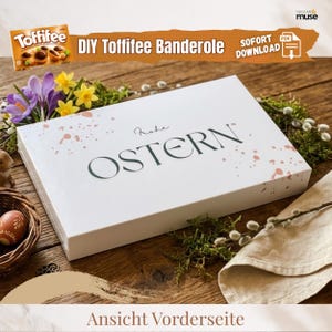May include: A white box with the word "OSTERN" in dark green lettering, decorated with pink paint splatters. The box is on a wooden surface with flowers and greenery. A Toffifee box is in the upper left corner. Text reads "DIY Toffifee Banderole" and "SOFORT DOWNLOAD".
