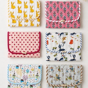 Könnte beinhalten: Sechs rechteckige, gesteppte Stofftaschen mit Klappverschlüssen, die durch Holzknebel gesichert sind. Die Designs umfassen gelbe Kaninchen auf Weiß, florale Muster in Pink, rosa Herzen, Rehe im Wald, eine bunte Dorflandschaft und kleine Tierfiguren.