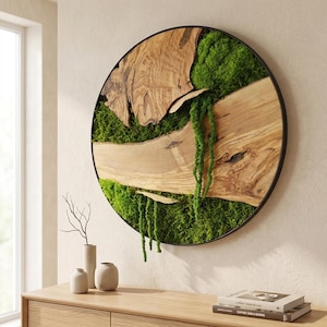 May include: A circular wall art piece featuring a dark metal frame enclosing natural wood slices with prominent grain patterns and vibrant green preserved moss. The moss includes hanging tendrils that drape over the wood surface.