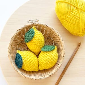 Puede incluir: Tres llaveros de limón hechos a mano con ganchillo, con hojas verdes y anillos plateados, están en una cesta de mimbre. Una madeja de lana amarilla y un ganchillo de madera también están en la imagen. Los limones son amarillos con una hoja verde.