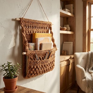 May include: A handmade, brown crochet wall organizer with wooden dowels and a hanging cord. The organizer is filled with books, notebooks, and pens. The word "CHRISTINA" is visible on one of the books. The wall is a neutral color.