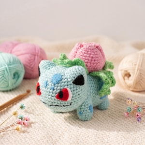 May include: A crocheted Bulbasaur figure in light blue, with a pink flower on its back and green leaves. The figure has black eyes, red accents, and blue spots. The background includes yarn balls and a crochet hook.