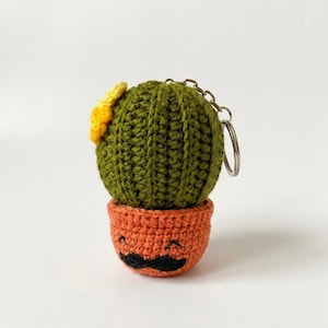 May include: A crocheted cactus keychain. The cactus is green with a yellow flower and sits in an orange pot with a smiling face and a black mustache. A silver chain and keyring are attached.
