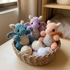 Puede incluir: Tres dragones de ganchillo hechos a mano en una cesta de mimbre. Los dragones son de color azul, lila y melocotón, con vientres y alas grises. La cesta también contiene ovillos de lana de varios colores.