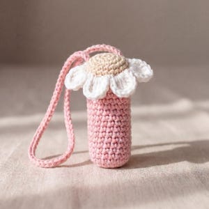 Puede incluir: Un portabotellas de agua hecho a mano con ganchillo en un tono rosa suave. El soporte es cilíndrico con un diseño de flor de margarita en la parte superior, con pétalos blancos y un centro beige. Se adjunta una correa rosa a juego para llevarlo. El artículo está hecho de hilo.