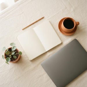May include: A closed dark gray laptop, an open blank notebook, a wooden pencil, a terracotta cup filled with black coffee on a matching saucer, and a small green plant in a terracotta pot arranged on a textured off-white fabric surface.