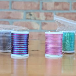 May include: Eight cylindrical spools of thread in various colors including blue, orange, grey, purple, pink, green, and red. Each spool is wrapped in a clear plastic bag and features a white plastic base and top, arranged in a horizontal row.