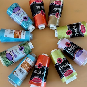May include: A collection of ten cylindrical spools of Maxi-Lock and Toldi-Lock serger thread arranged in a circle. The spools feature various solid colors including orange, tan, light green, pink, and blue, along with two variegated swirl patterns.