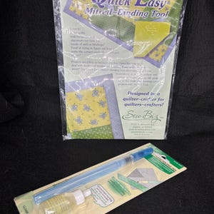 May include: A Quick Easy Mitred-Binding Tool in a clear plastic package with purple and yellow floral fabric graphics, and a Clover Fabric Folding Pen set including a blue plastic pen and a small bottle of concentrated marking solution in packaging.