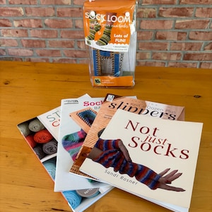 May include: A clear plastic package containing a blue sock loom with the text Sock Loom Knit your own socks, and a stack of four instructional books with titles including Slippers and Not just Socks, featuring colorful yarn and knitted projects.