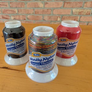 May include: Three spools of YLI Woolly Nylon serger thread on white plastic bases. One spool is black, one is red, and the center spool features a multicolored variegated pattern. Each label reads Woolly Nylon Serger Thread for knits and edges.