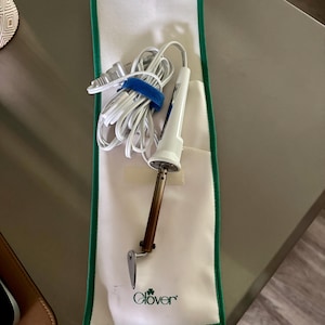 May include: A Clover brand electric fabric tool with a white handle, a metallic heating element, and a pointed tip. The tool includes a white power cord with a plug and is resting on a white fabric storage case with green trim and the Clover logo.