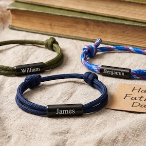 Könnte beinhalten: Drei personalisierte Seilarmbänder in Grün, Blau und Blau-Rot, jedes mit einem schwarzen Metallstab, auf dem ein Name eingraviert ist. Eine Karte mit "Happy Father's Day" ist ebenfalls vorhanden, zusammen mit einem Stapel alter Bücher.