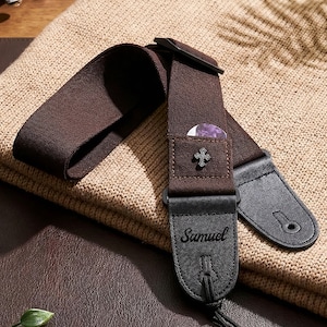 以下が含まれることがあります： 茶色のギターストラップ。黒い革製の端があり、「Samuel」という名前がプリントされています。紫色のピックと装飾的な十字架が入った小さなポケットが付いています。ベージュ色のニットセーターの上に置かれています。