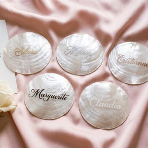 May include: Five personalised seashell place cards, each inscribed with a name in elegant script. The names "Silvia", "Manny", "Cristianna", "Marguerite", and "Claudia" are displayed on pearlescent shells, on a pink satin backdrop.