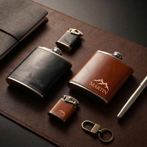 Könnte beinhalten: Eine Auswahl an personalisierten Lederflaschen, Feuerzeugen und Accessoires. Die Flaschen sind schwarz und braun, mit silbernen Akzenten. Eine Flasche ist mit dem Namen "Matthew" graviert, eine andere mit "MARTIN" und einer Berg-Grafik. Ein Stift, ein Notizbuch und ein Schlüsselanhänger sind ebenfalls vorhanden.