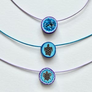 Puede incluir: Tres collares con colgantes redondos con diseños de vida marina. El colgante superior tiene un diseño de orca azul y blanco, el colgante central tiene una tortuga y el colgante inferior tiene un diseño de tortuga. Los cordones son azules y morados.