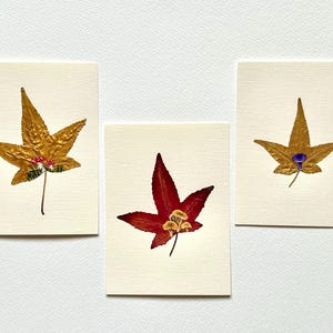 May include: Three botanical art pieces featuring pressed leaves. One gold leaf with red accents, a red leaf with tan accents, and a gold leaf with a blue accent. Each leaf is mounted on a cream-colored card.