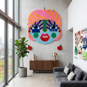 May include: A large, colorful, tufted wall hanging depicting a stylized face with pink hair, blue skin, and red lips. Two small red apple-shaped ornaments hang from the bottom. The piece features bold geometric patterns and vibrant yarn textures.