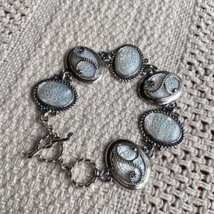 May include: A silver-toned bracelet featuring five oval links with white, patterned, iridescent inserts. Each oval link is framed by a twisted rope-style border and accented with a silver swirl design. The bracelet includes a toggle clasp closure.
