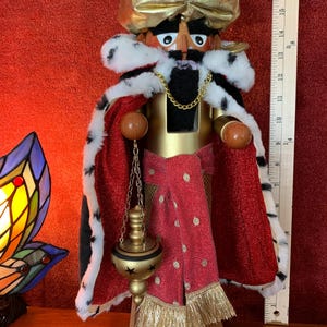 May include: A decorative nutcracker figurine of Balthasar, featuring a gold crown, red cape with white fur trim, and a gold chain. The figurine has a black beard and holds a gold censer. The base reads "C+M+B BALTHASAR C+M+B".