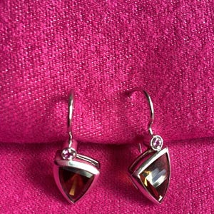 May include: A pair of silver drop earrings featuring triangular-cut dark amber gemstones. Each earring is accented with a small, round, pink gemstone set at the top of the hook, creating a distinct contrast against the deep brown of the main stone.