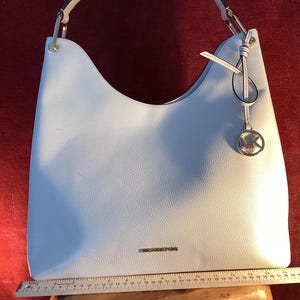 May include: A light blue Michael Kors shoulder bag with a single handle and a silver-tone MK logo charm. The bag has a textured finish and a rectangular shape. The bag is approximately 15 inches wide.