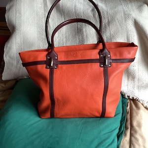 May include: An orange leather tote bag with dark brown accents and handles. The bag has a rectangular shape with a structured design, featuring vertical brown stripes and gold-colored hardware. The handles are rounded and also dark brown.
